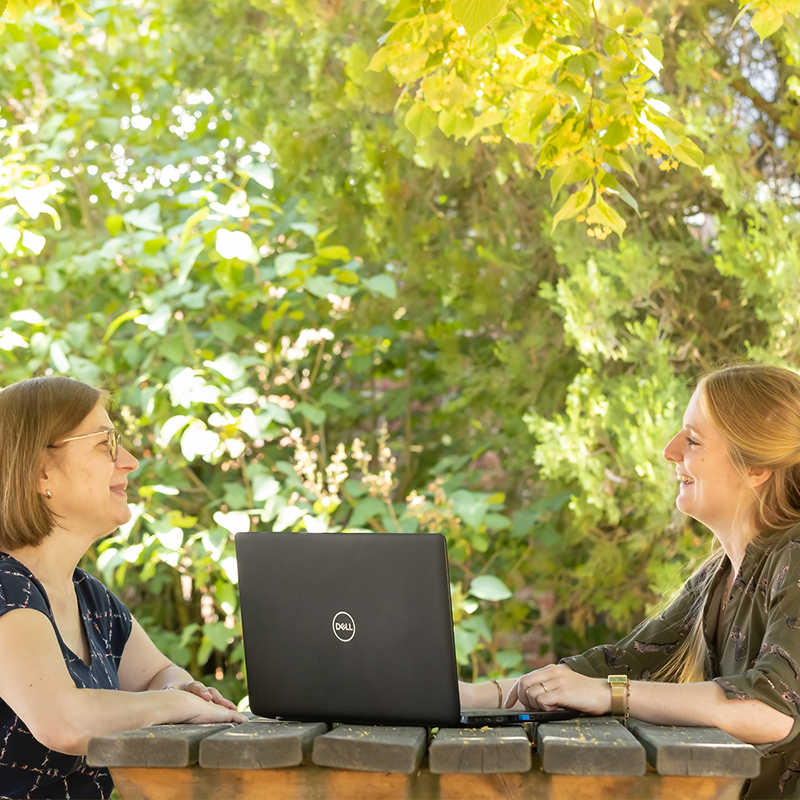 Twee personen zijn in gesprek aan een picknicktafel met een laptop tussen hen in